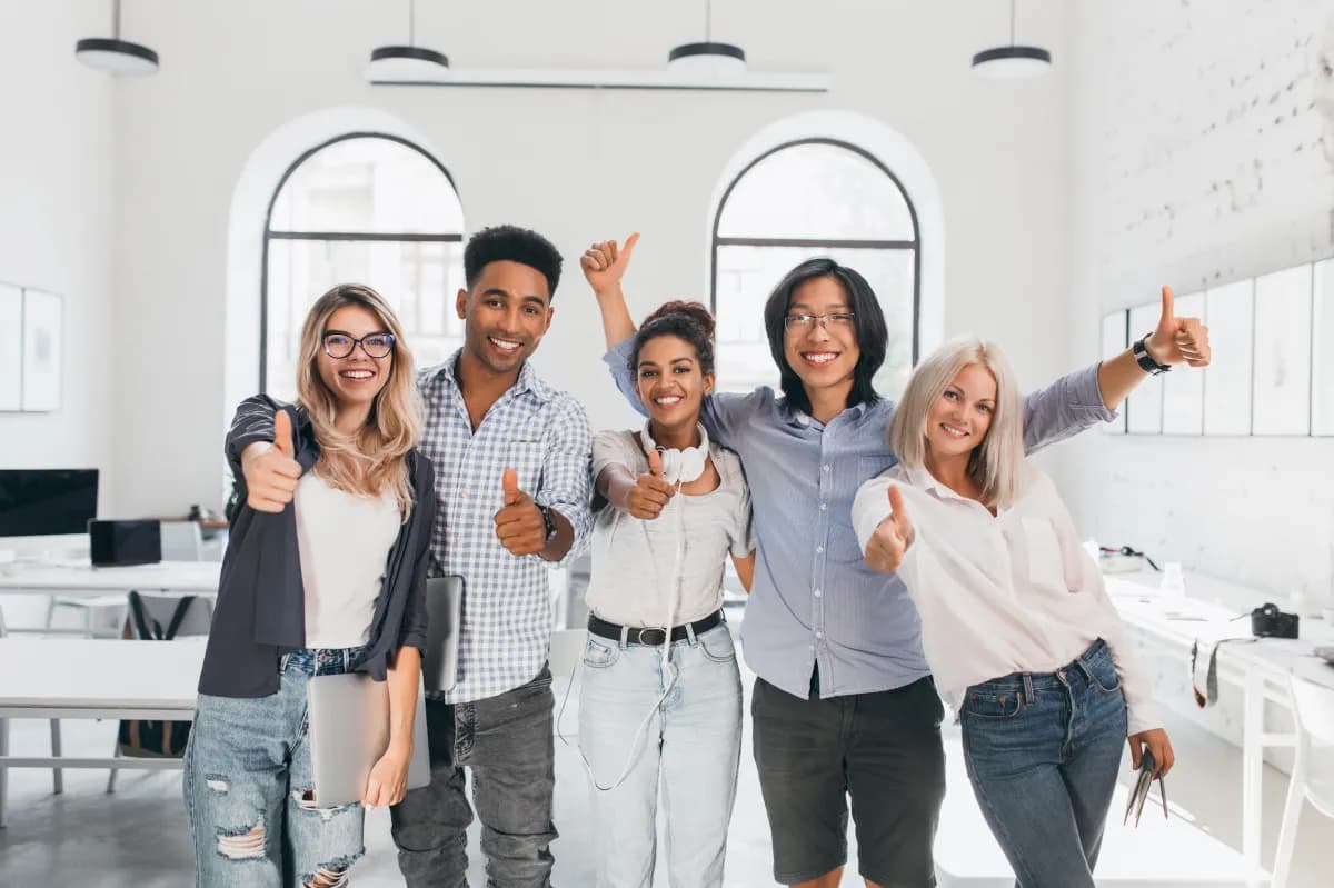 Five diverse coworkers standing in an office space, smiling and giving thumbs up, expressing teamwork and positivity.